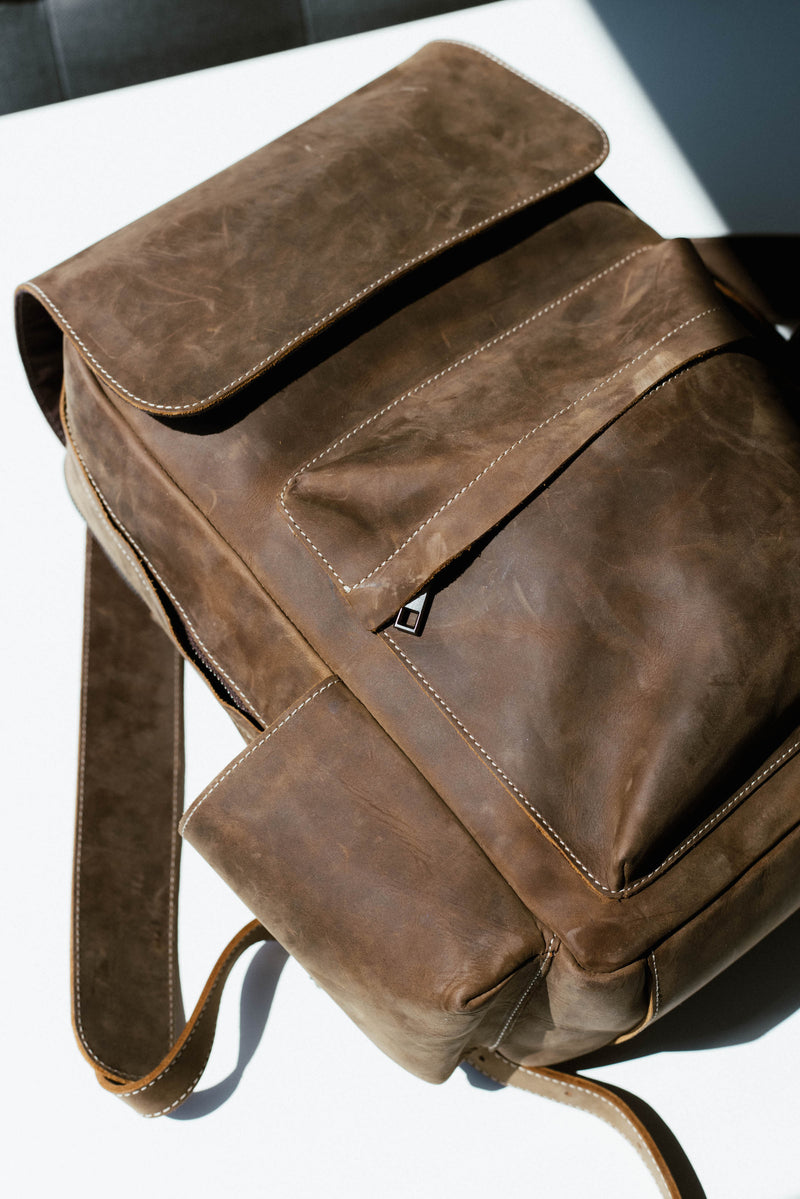 Brown leather backpack with white stitching and zipper pull, photographed close-up on a sunlit white surface.