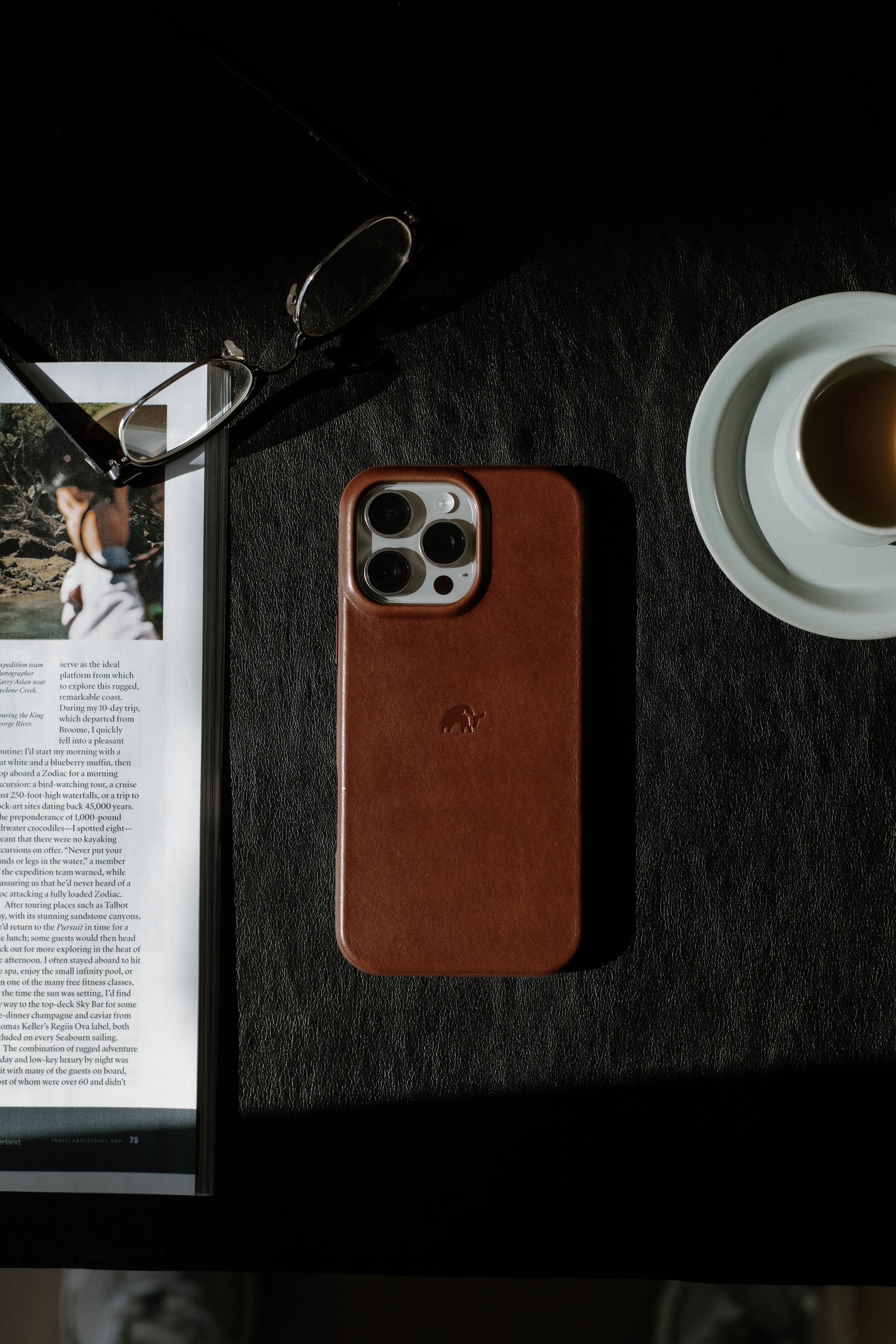 A brown leather phone case on a table next to a magazine and a cup of coffee.