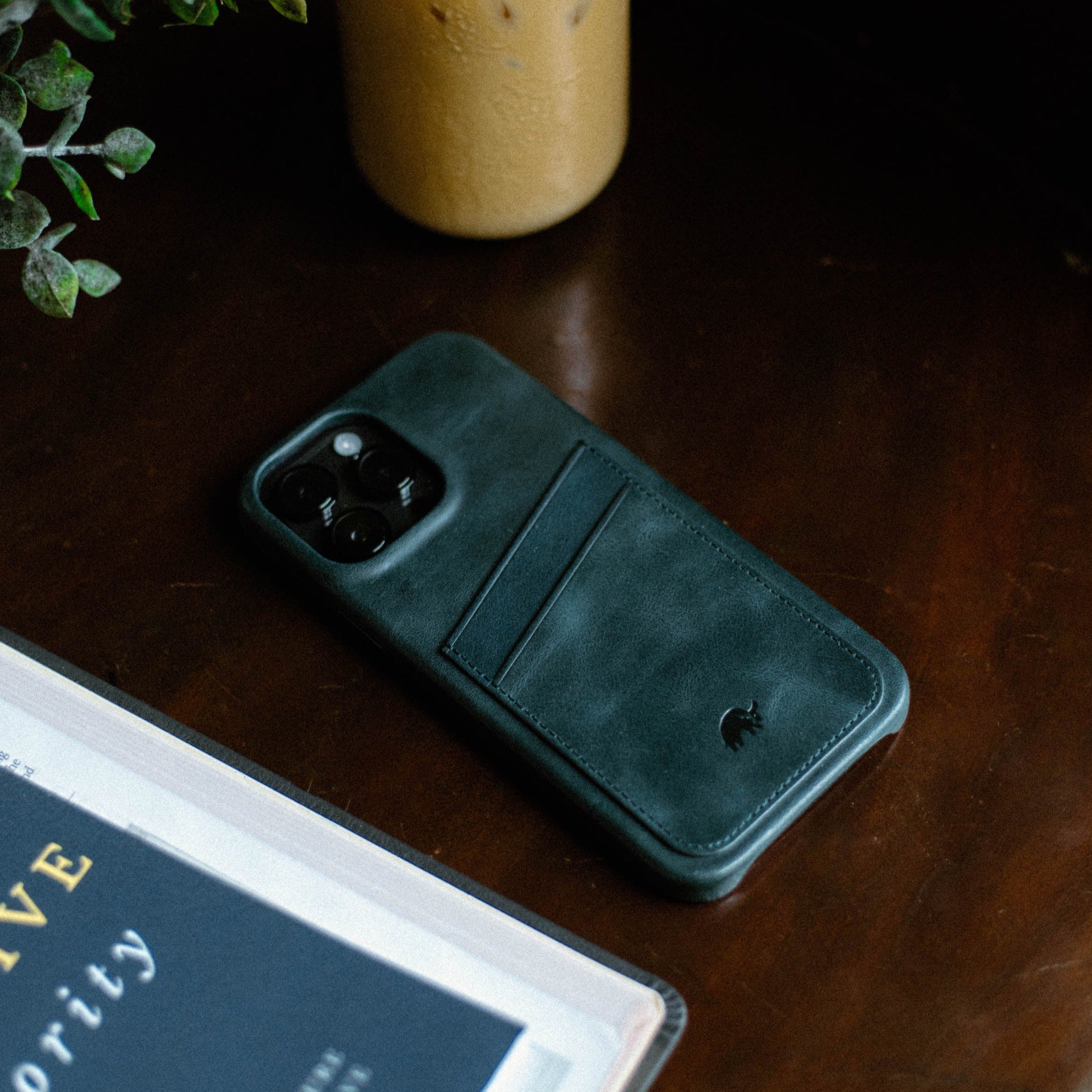 Dark green leather iPhone case with card pocket on wooden table; book corner shows text 'VE' and 'ority'.