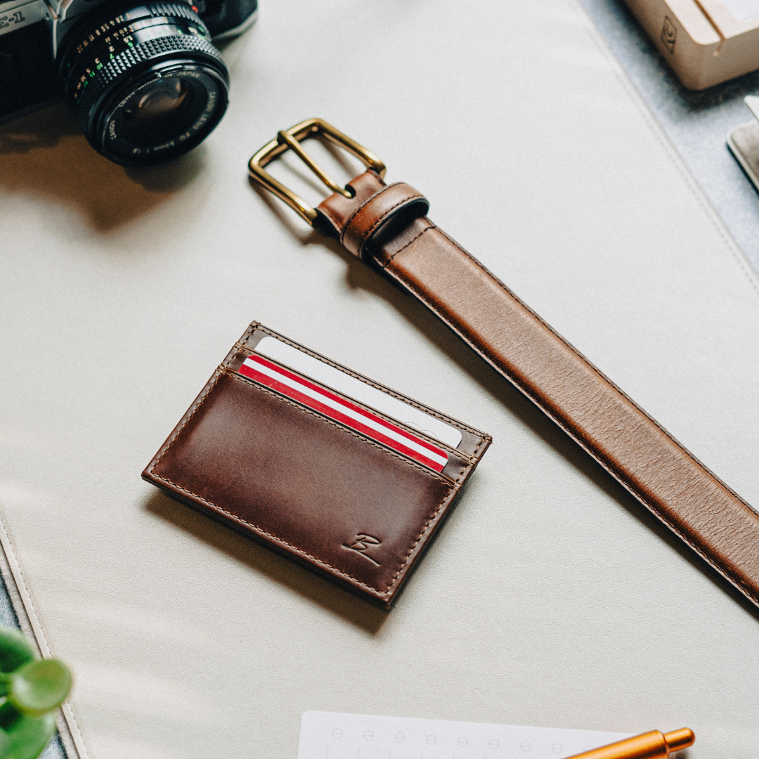 Brown leather cardholder with cards on a desk next to a belt and camera