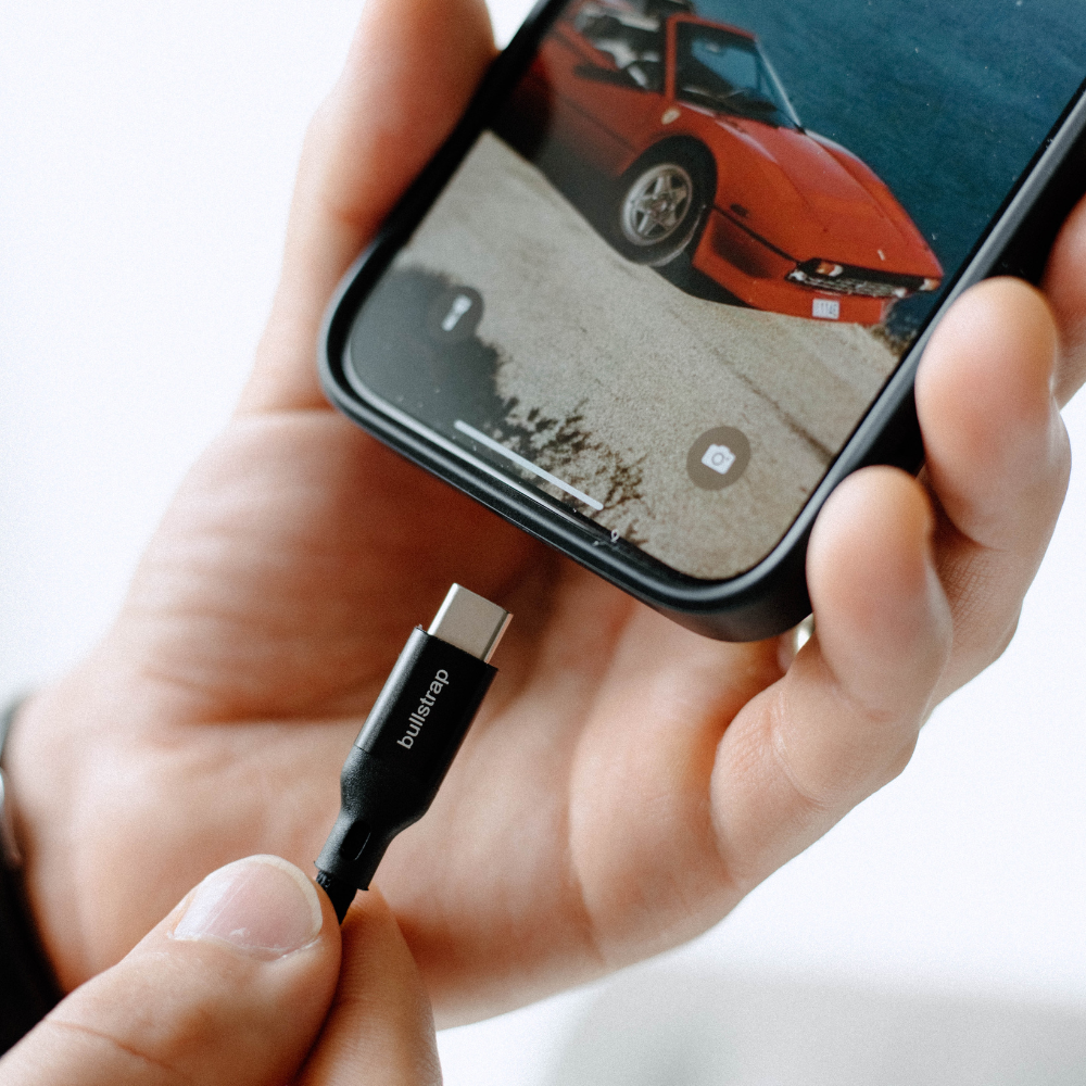 Hand holding a smartphone as a USB-C cable labeled bullstrap is held near the phone's charging port.