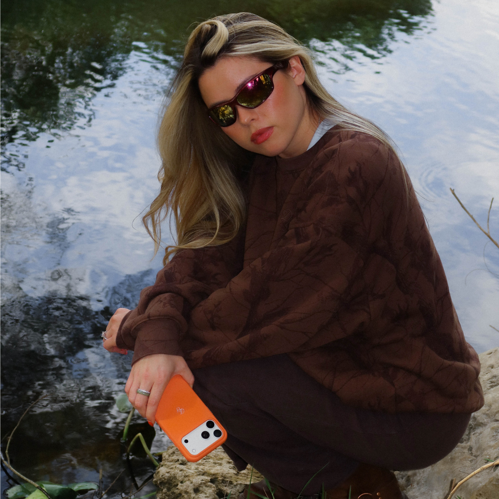 Young woman crouching by a pond holding a smartphone with a Hunter Orange leather case