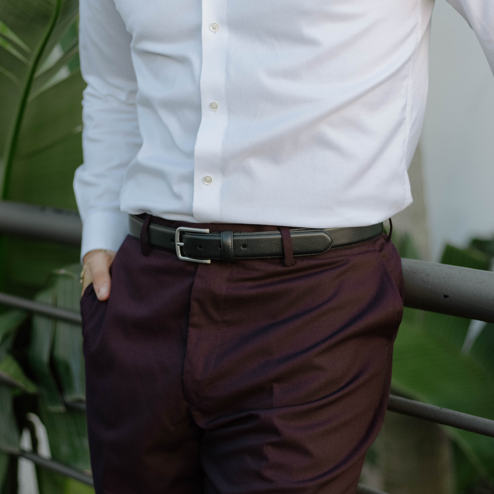 A person wearing a black belt with a white shirt and dark pants, standing near greenery.