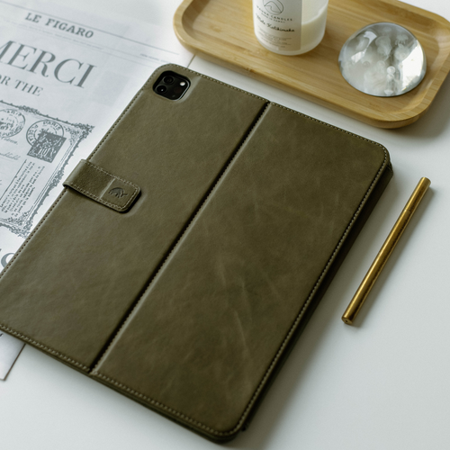 Olive green leather iPad case with a flap and embossed logo on a white background with a pen and decorative items.