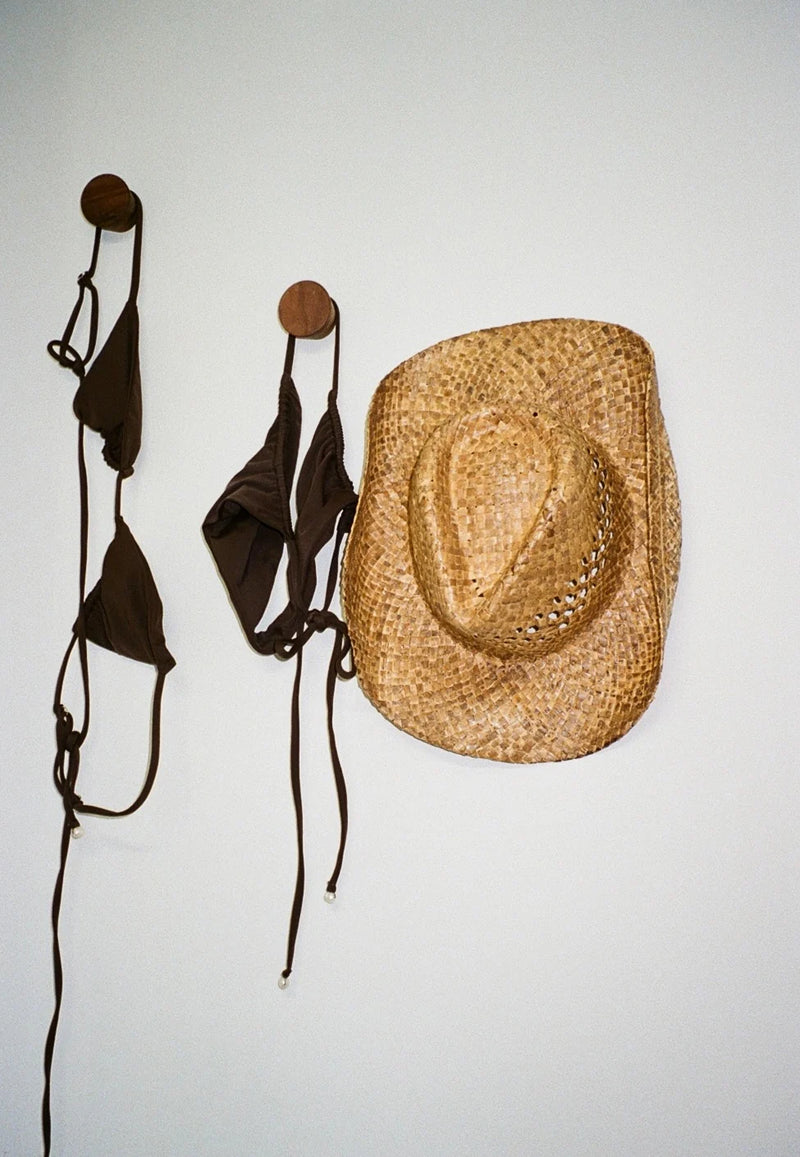 Straw cowboy hat and brown bikini tops hanging on wooden pegs against a white wall.