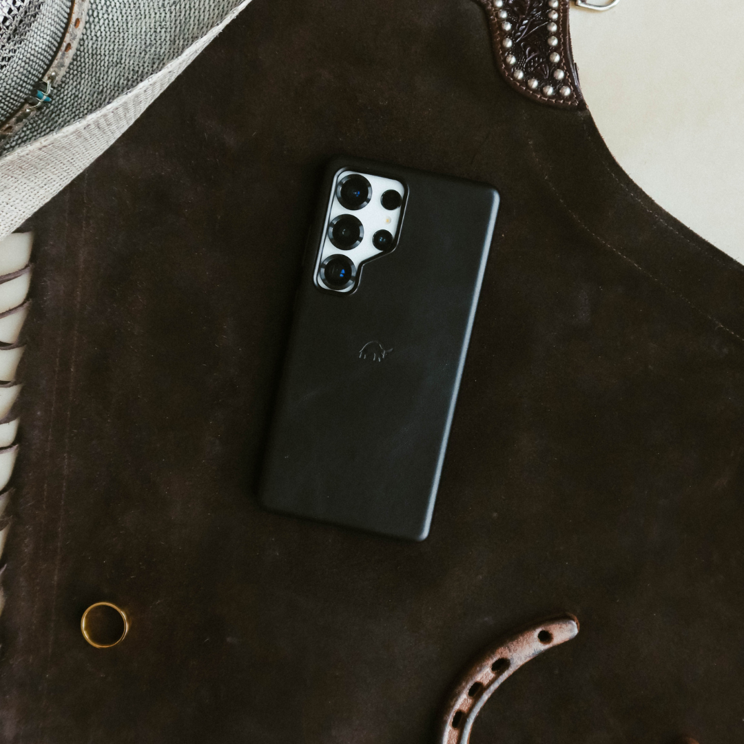 Black smartphone with four rear cameras on dark brown leather, beside a gold ring and a leather strap.