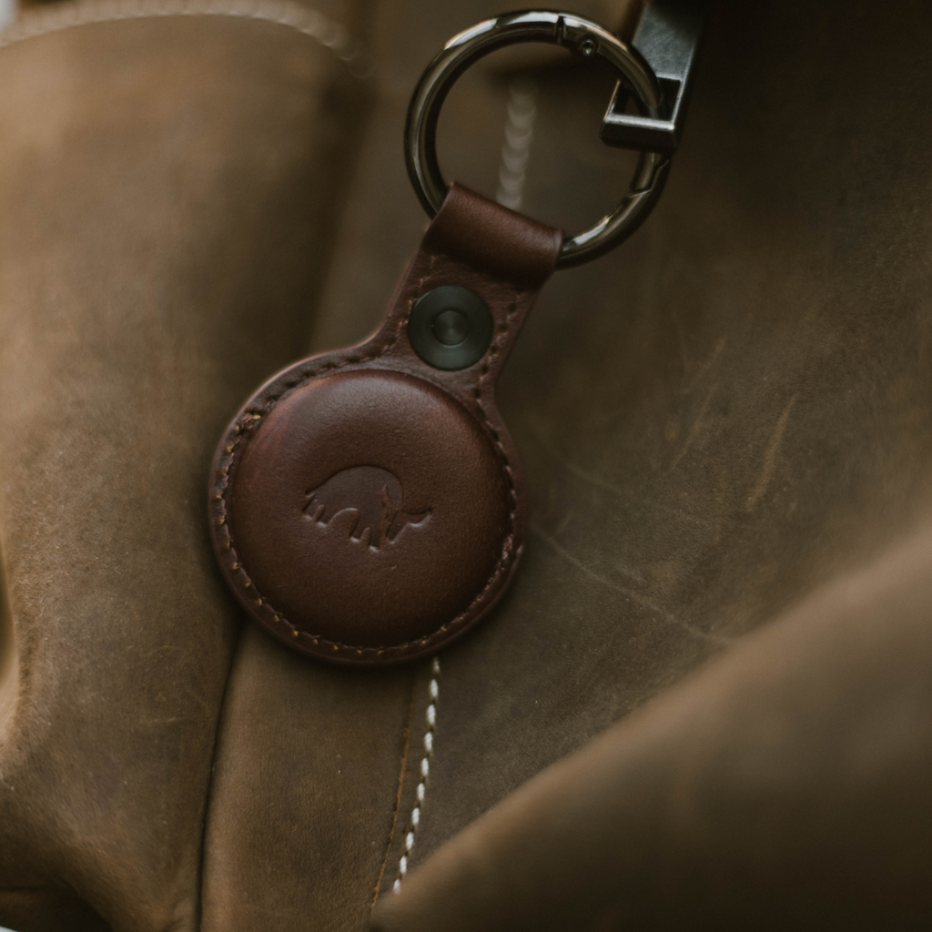 Brown leather key fob with embossed elephant logo attached to a metal key ring, resting on a stitched leather bag.