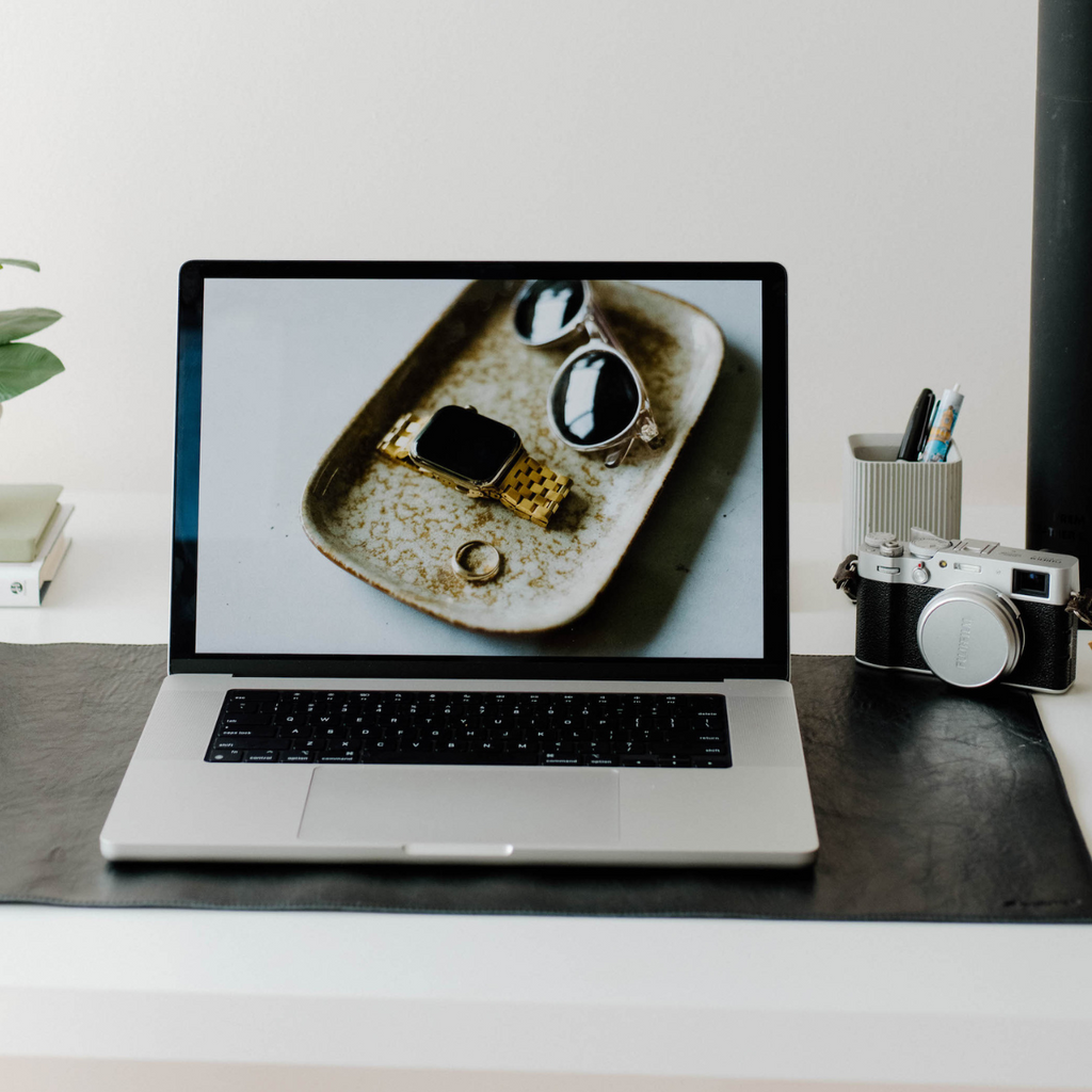 Laptop on black leather desk mat with Fujifilm camera at right; screen shows sunglasses, gold watch, and ring.