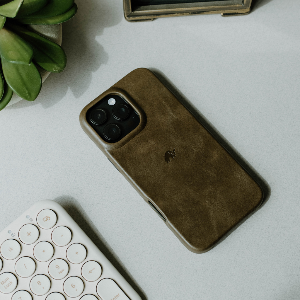 Brown leather phone case on a light desk beside a white round-key keyboard and a small succulent
