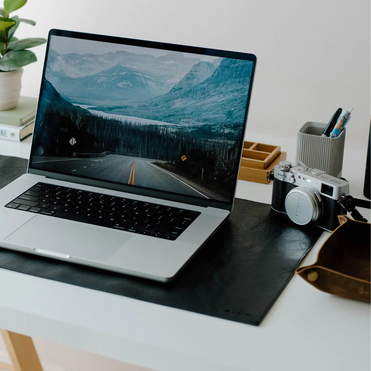 Black leather desk mat with a laptop and a camera on a white desk, plant and pen cup in background.