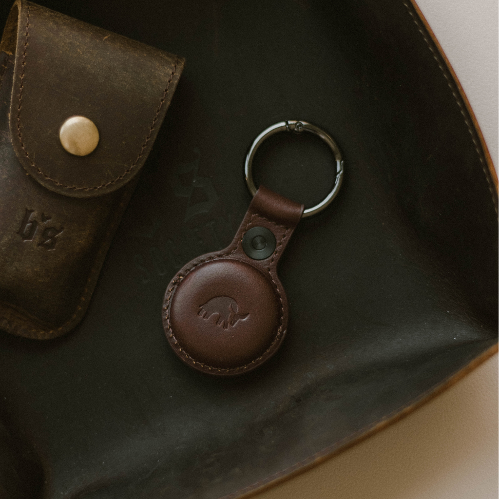 Top-down view of BullTag leather AirTag keyring showcasing embossed bull logo, snap closure, and metal keyring on a dark leather tray