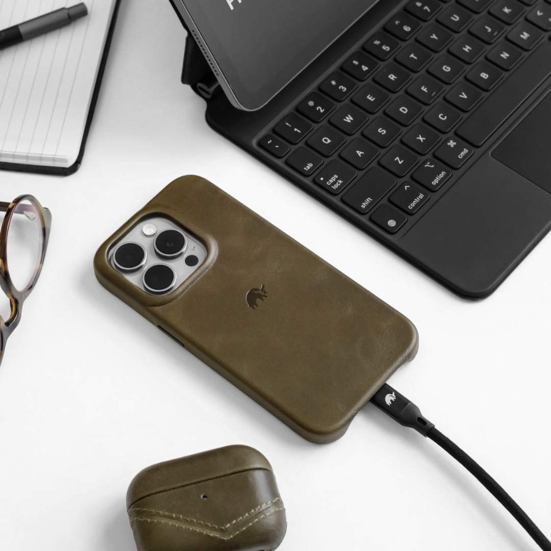 Olive leather iPhone case with elephant logo, charging cable connected, on desk beside keyboard, glasses, and earbuds case