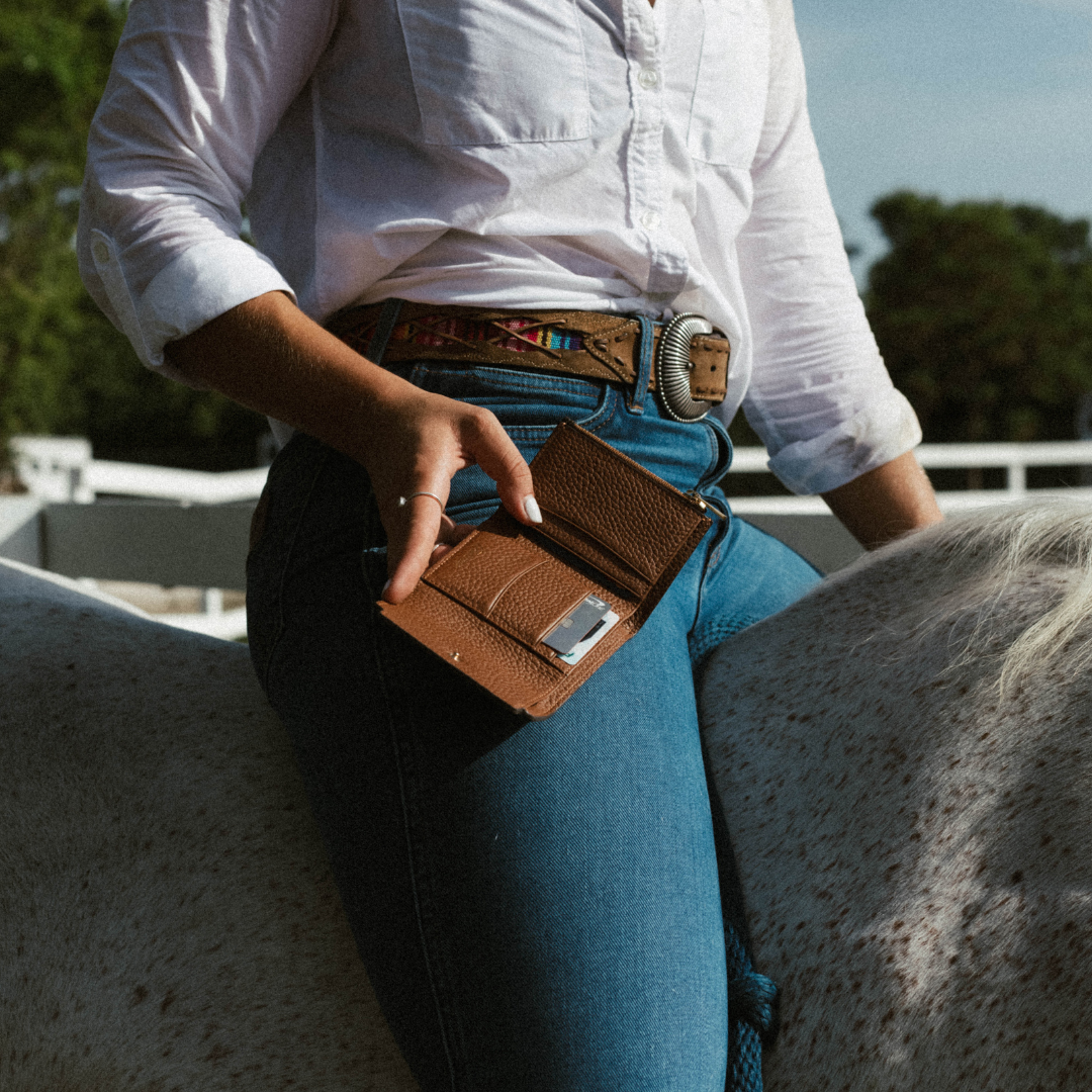 Rider in a white shirt and jeans seated on a speckled horse, holding an open brown leather wallet showing a card.