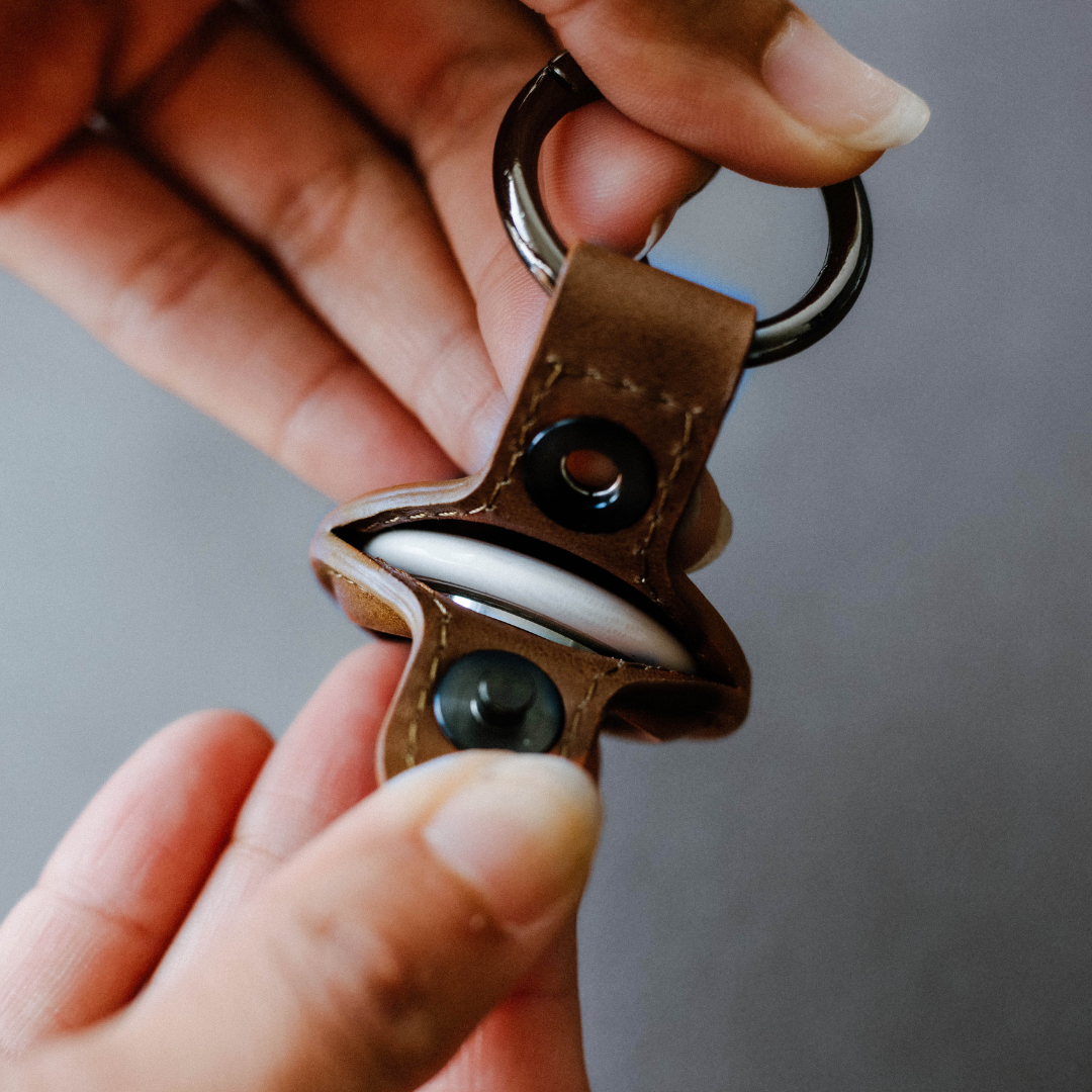 Brown leather BullTag keyring holding an AirTag, shown between two hands