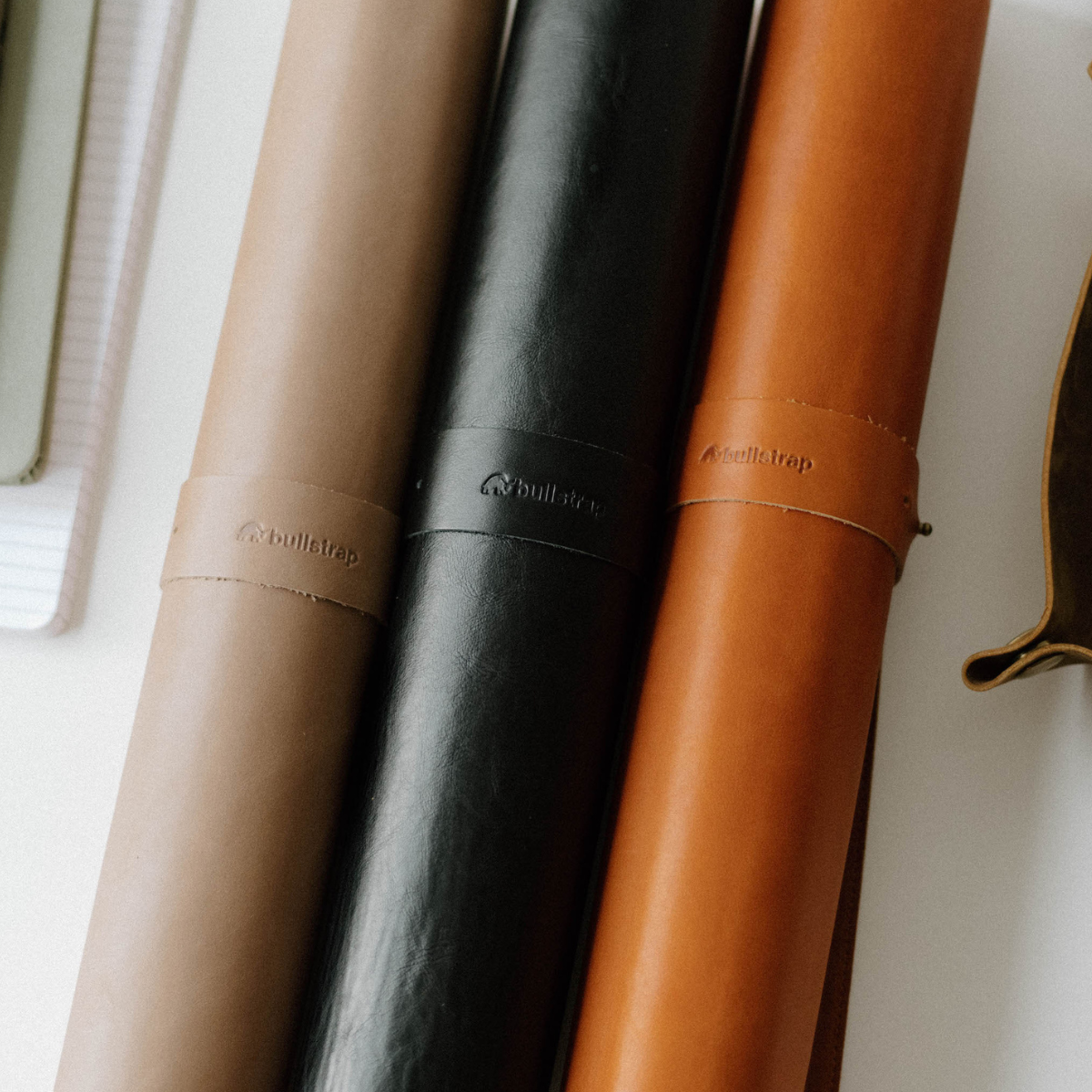 Three rolled leather desk mats—tan, black, and cognac—with embossed bullstrap straps.