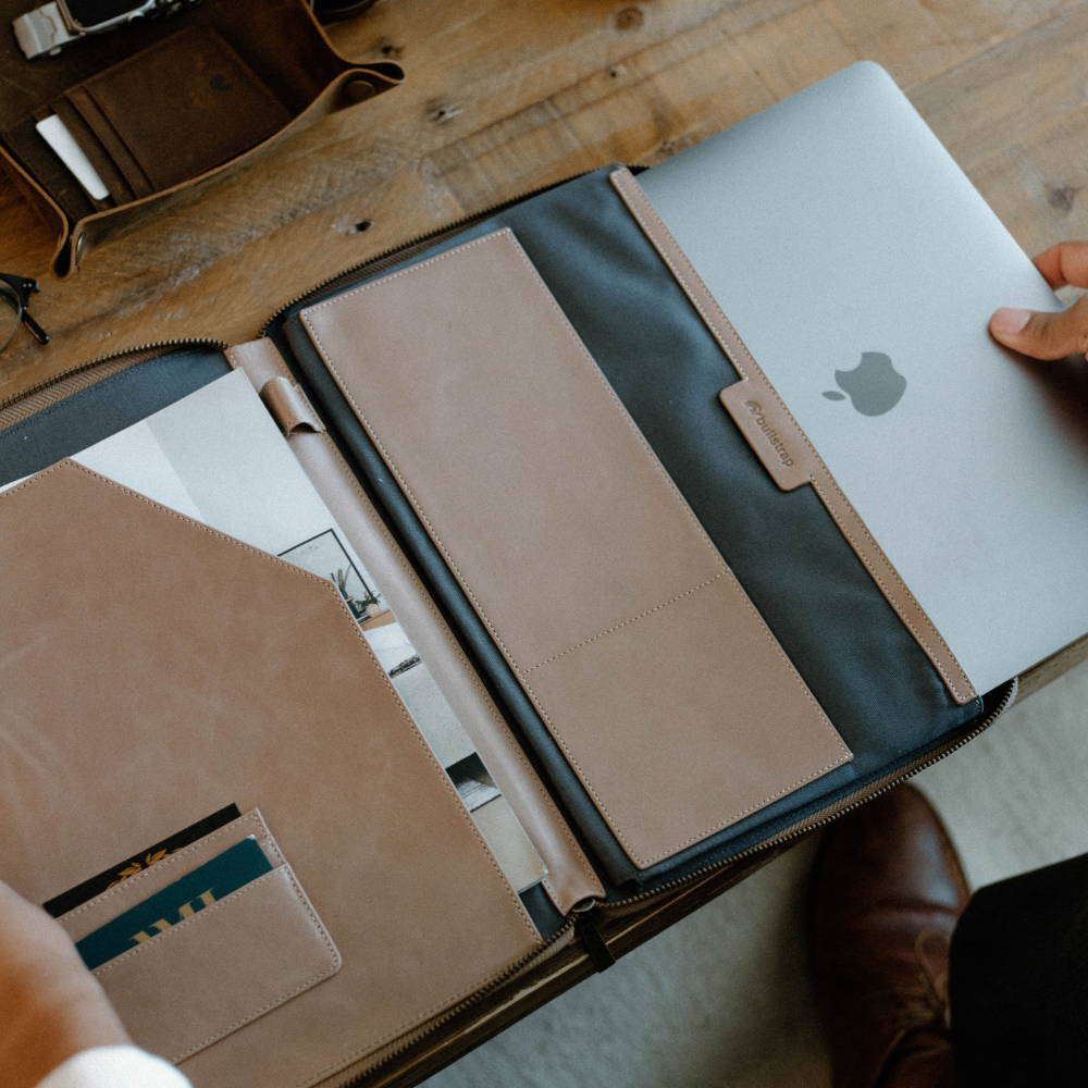 A person placing a MacBook into a leather sleeve with compartments for documents and cards.