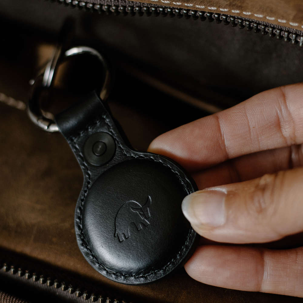 Black leather key fob with round tracker embossed with a mammoth logo, held between fingers beside a brown zipper.