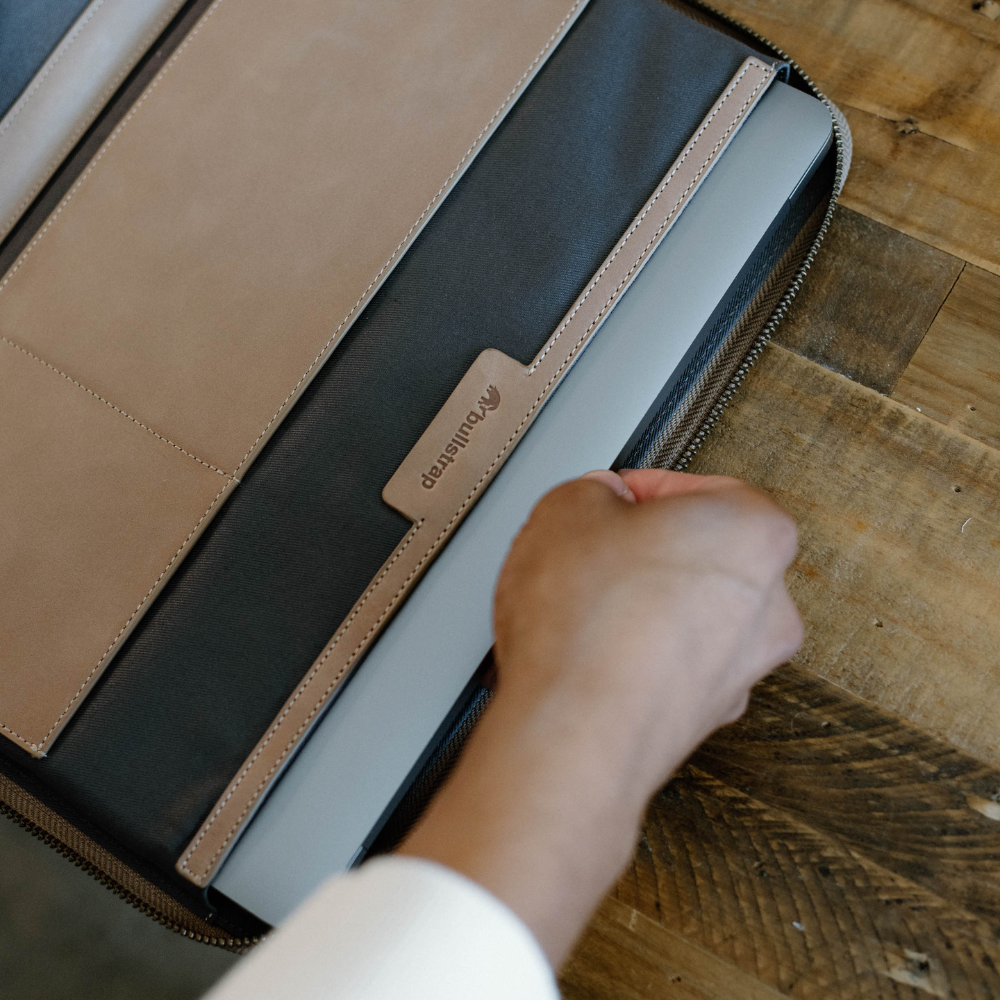 A hand holding a MacBook inside a leather sleeve on a wooden table.