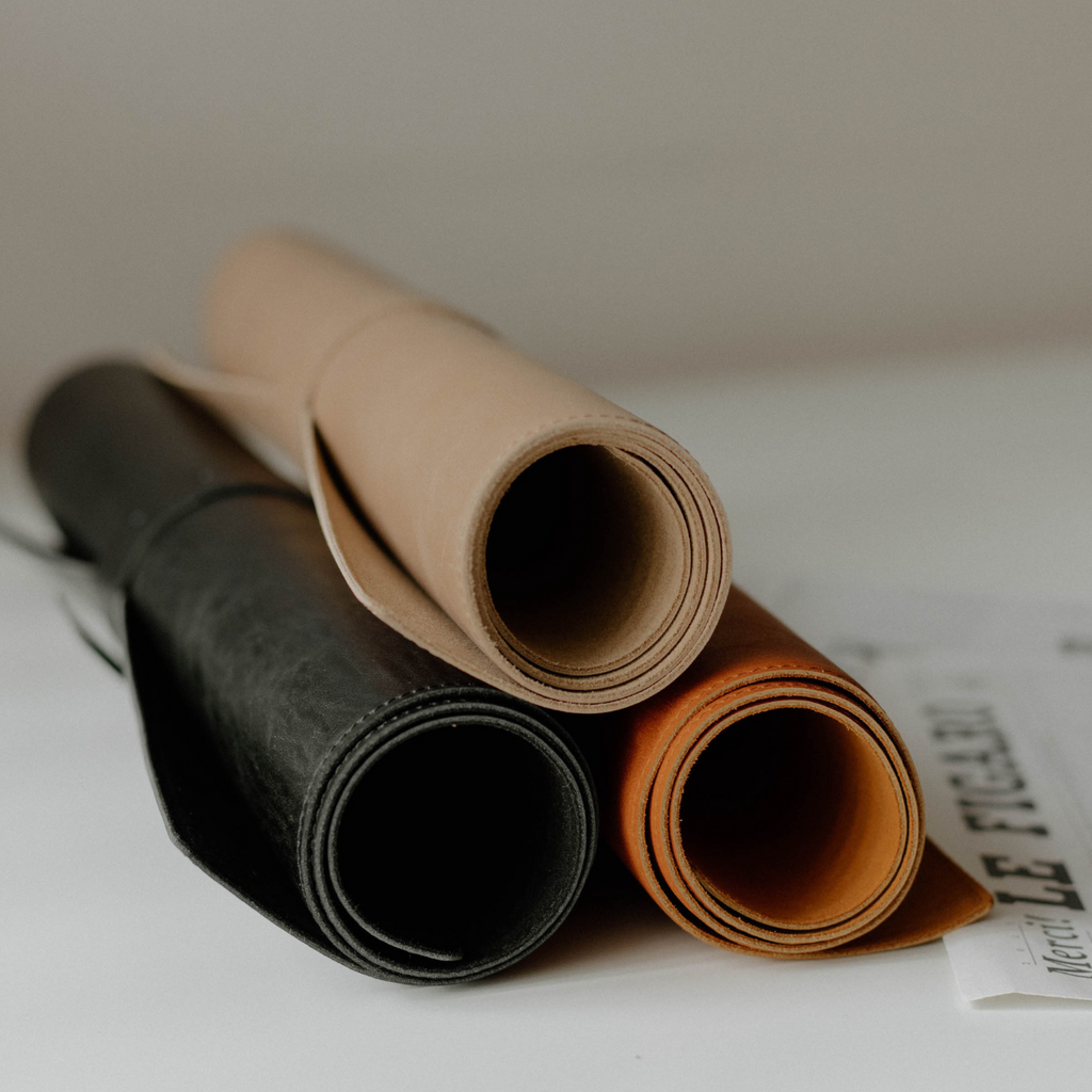 Three rolled leather desk mats in tan, black, and cognac stacked on a white surface
