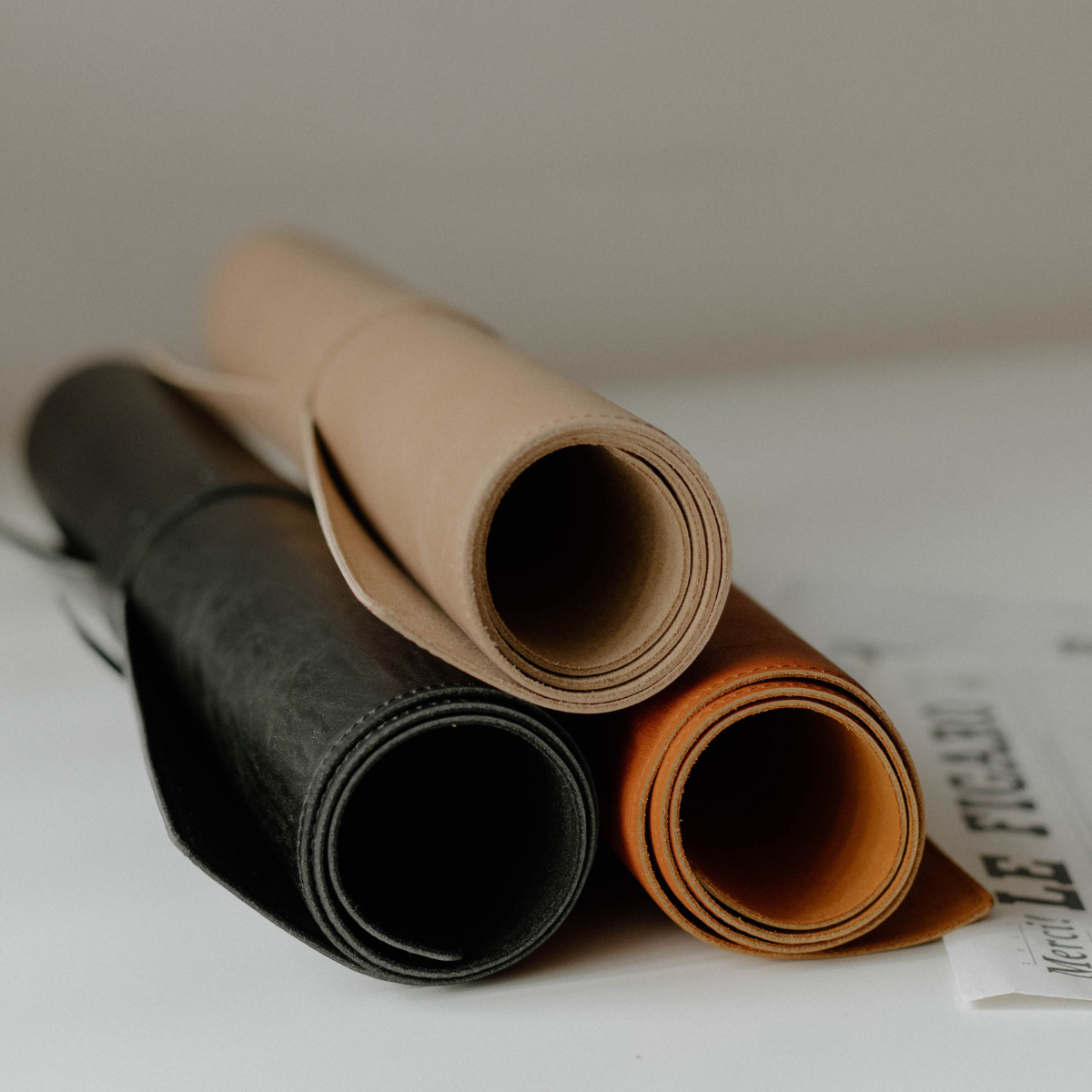 Three rolled leather desk mats in tan, black, and cognac stacked on a white surface
