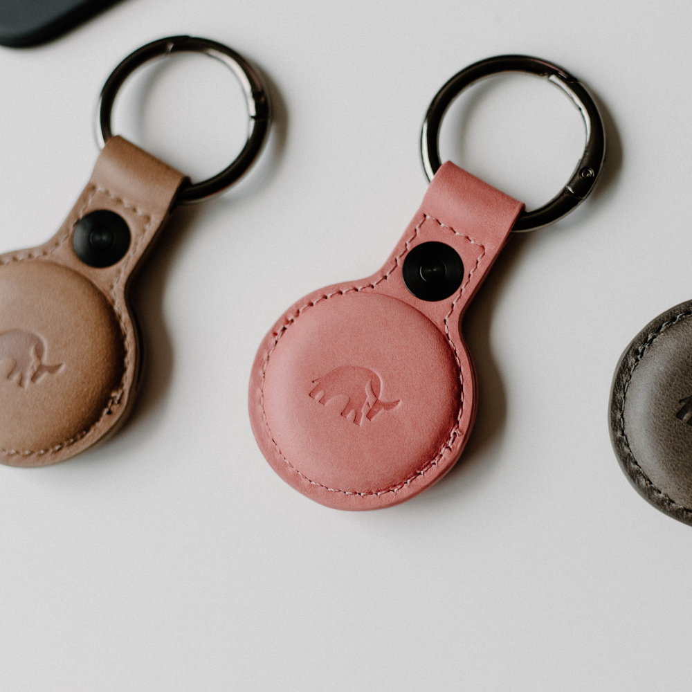 Three leather AirTag holders in various colors, including pink, displayed on a white surface.