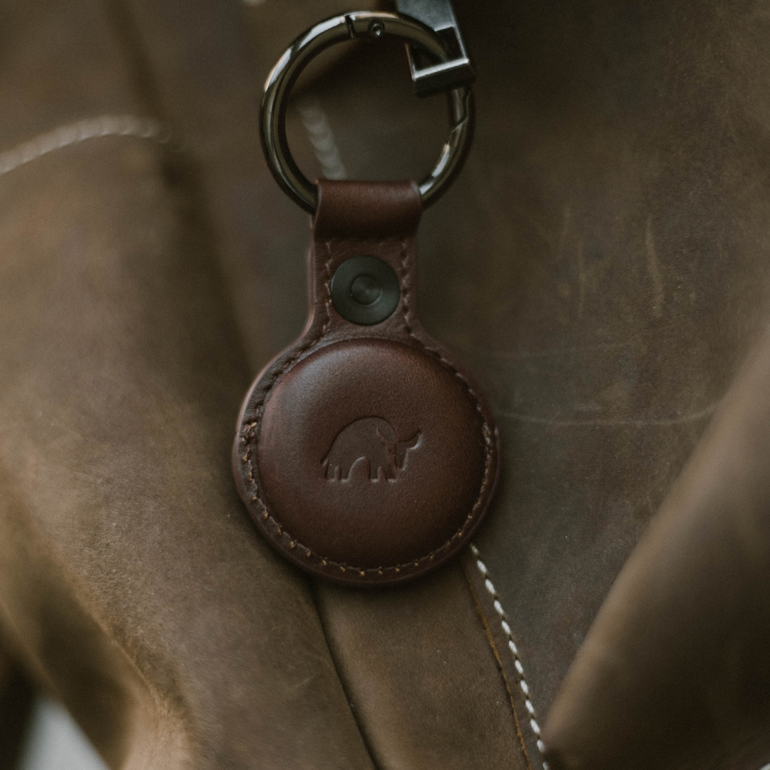 Brown leather AirTag keyring with embossed bull logo and metal split ring, attached to a waxed canvas bag.
