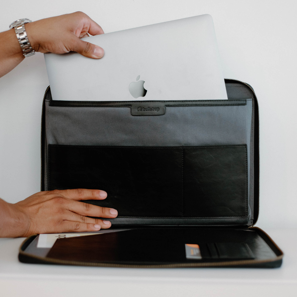 A person placing a MacBook into a leather laptop sleeve with compartments.