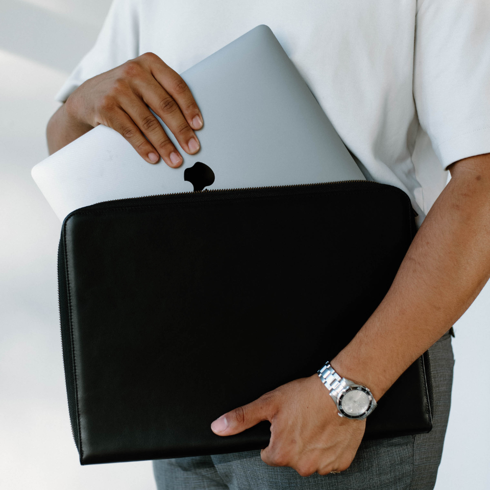 A person holding a black leather MacBook sleeve with a laptop inside.
