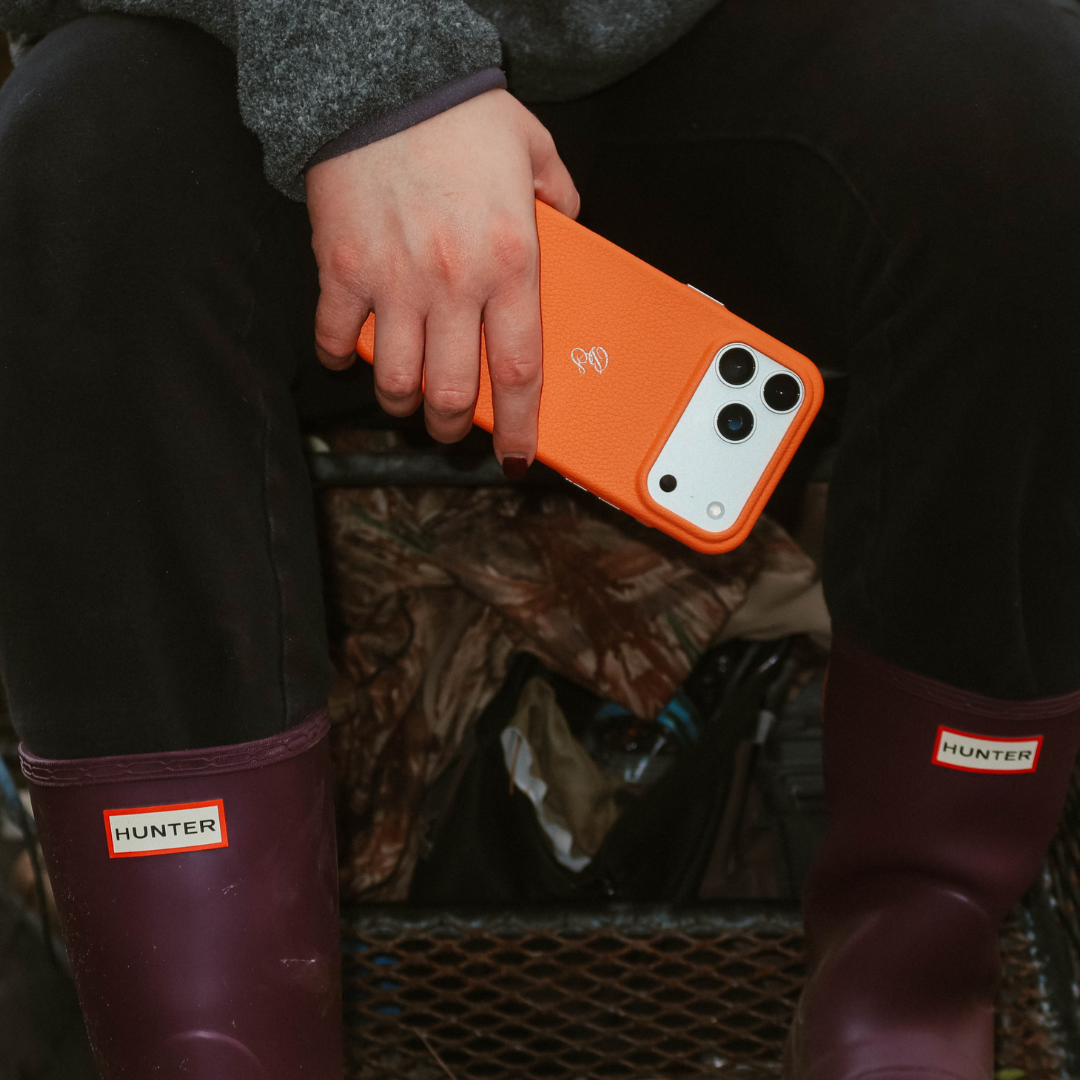 Hand holding iPhone in Blaze Hunter Orange leather case while seated between maroon Hunter boots and camo gear