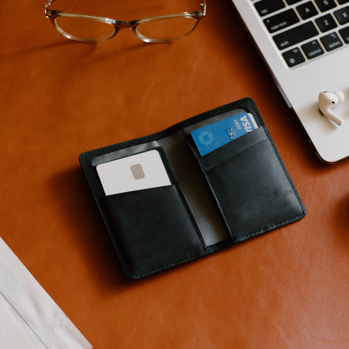 Open black leather bifold wallet on tan desk with two cards visible, one blue card labeled VISA; laptop corner and glasses.