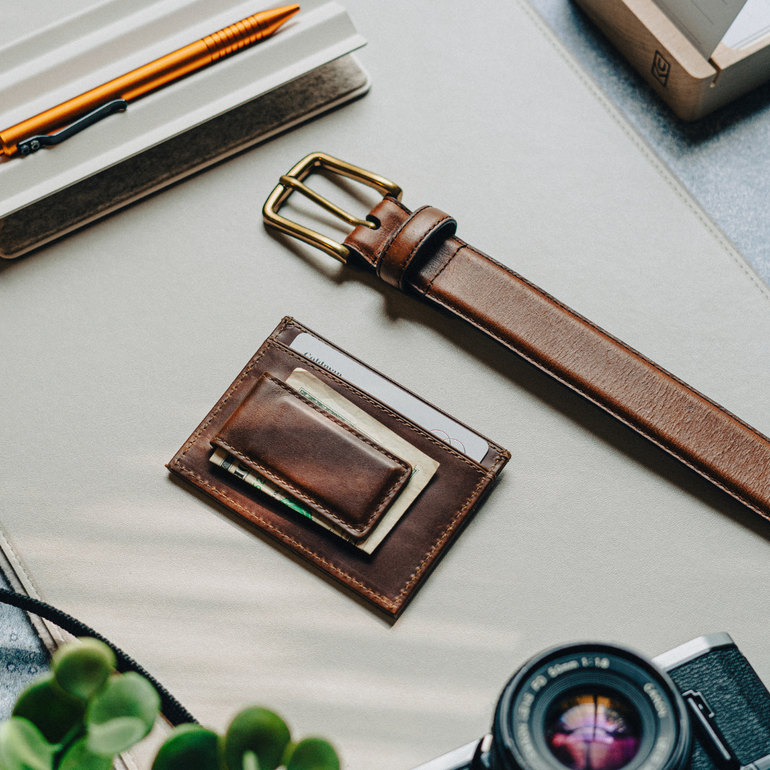 A brown leather money clip and card holder on a desk with a belt and camera.