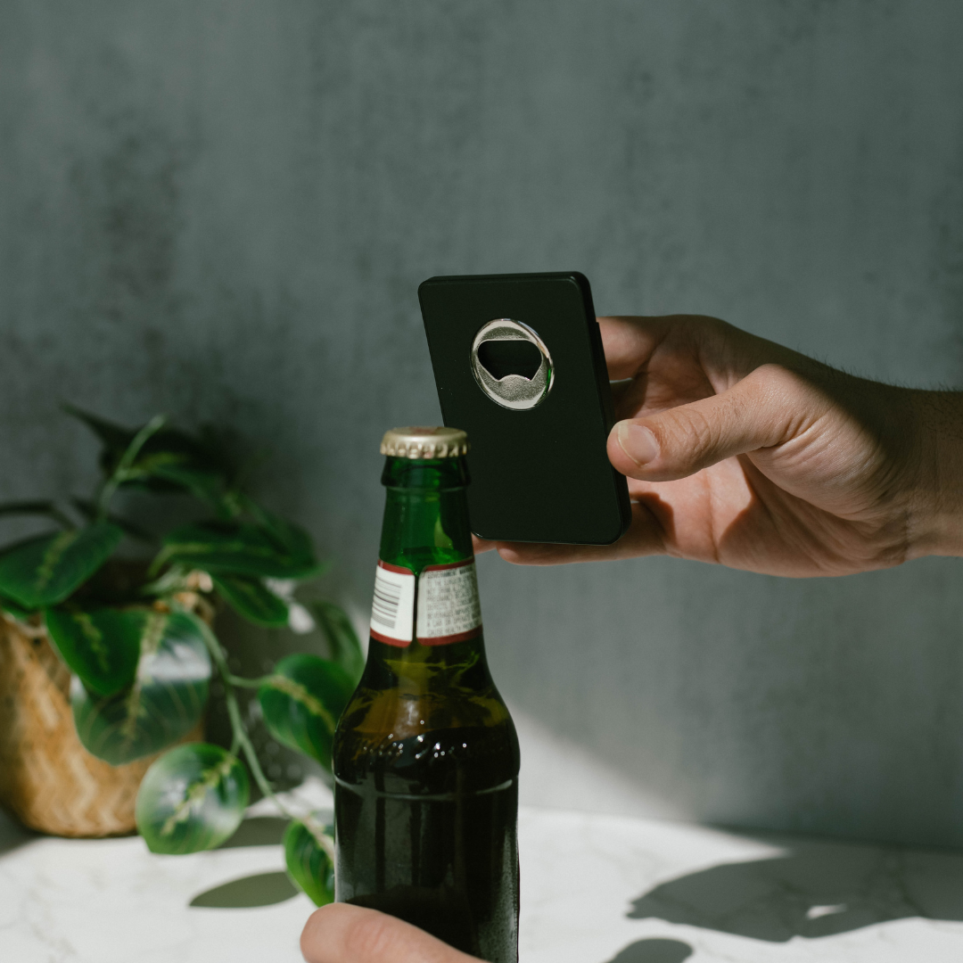 A hand holding a black Magnetic Popper Wallet with a built-in bottle opener, positioned above a beer bottle.