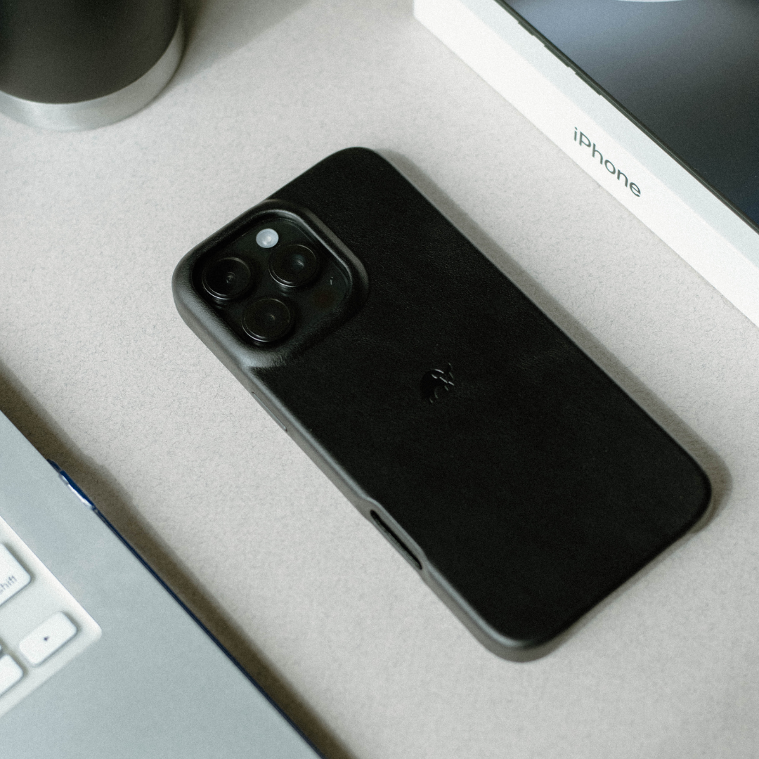 Black leather phone case with raised camera cutout and embossed logo, lying on a desk beside an iPhone box
