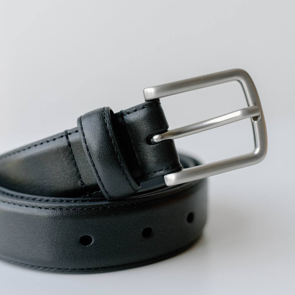 Black leather belt coiled with brushed silver rectangular buckle, close-up showing stitching and punched holes.