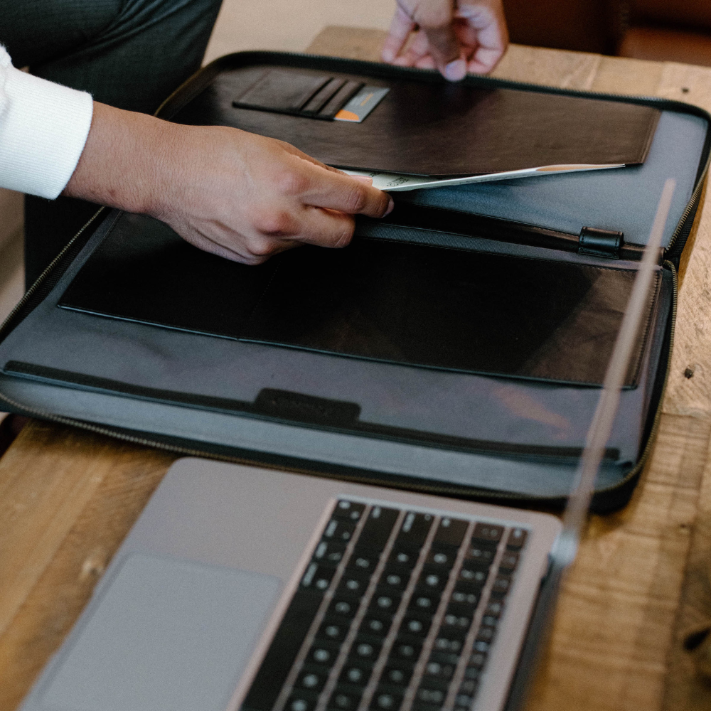 A person placing a document into a black leather MacBook sleeve on a wooden table next to a laptop.