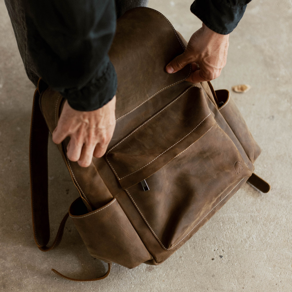 Hands opening a worn brown leather backpack on a concrete floor.