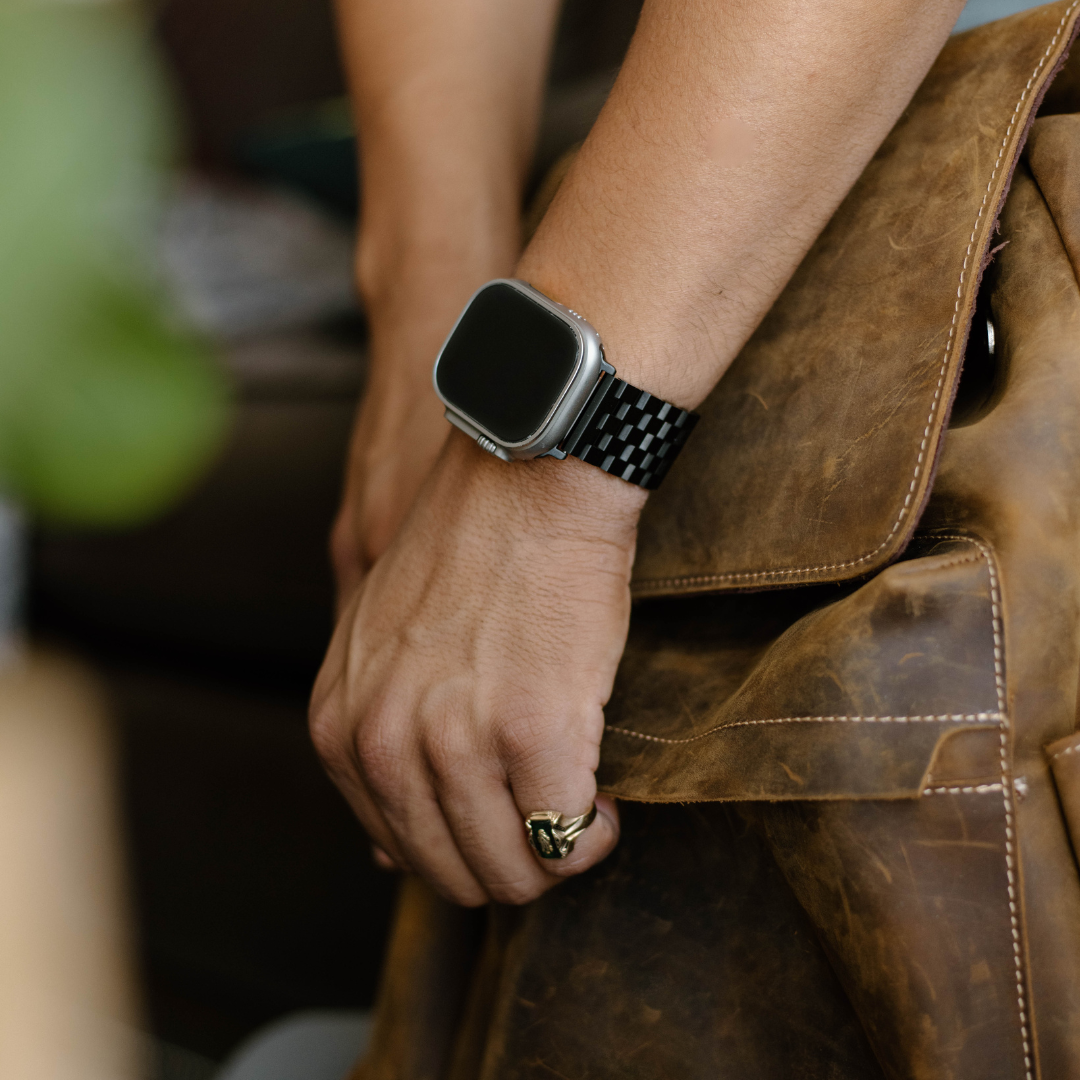 Black metal link Apple Watch strap shown on a wrist holding a leather bag