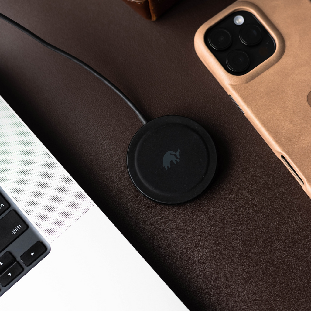 Black circular Qi2.0 MagSafe wireless charging puck with elephant logo on brown leather; nearby laptop edge shows 'shift'