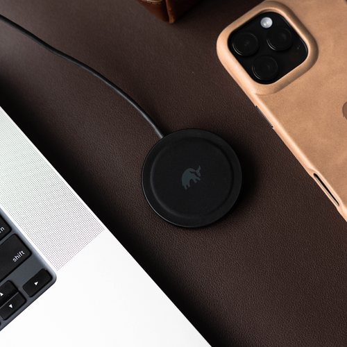 Black circular Qi2.0 MagSafe wireless charging puck with elephant logo on brown leather; nearby laptop edge shows 'shift'