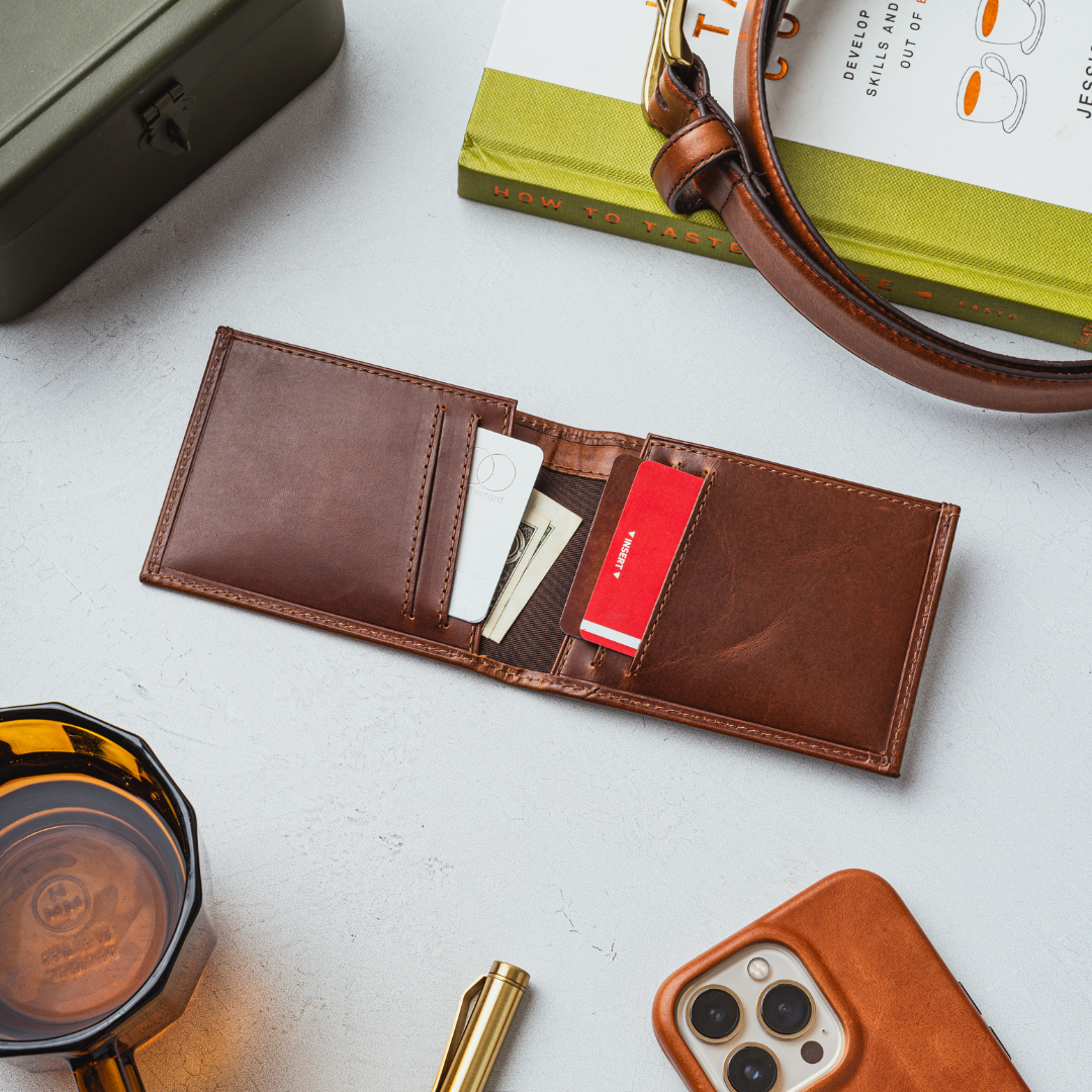 Brown leather bifold wallet open showing cards and cash on tabletop beside a book titled HOW TO TASTE and a drink
