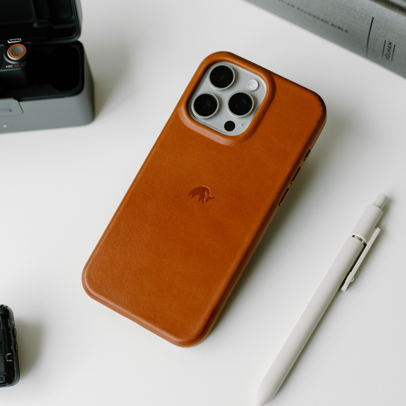 A brown leather iPhone case with a logo, placed on a white surface next to a pen and a camera.