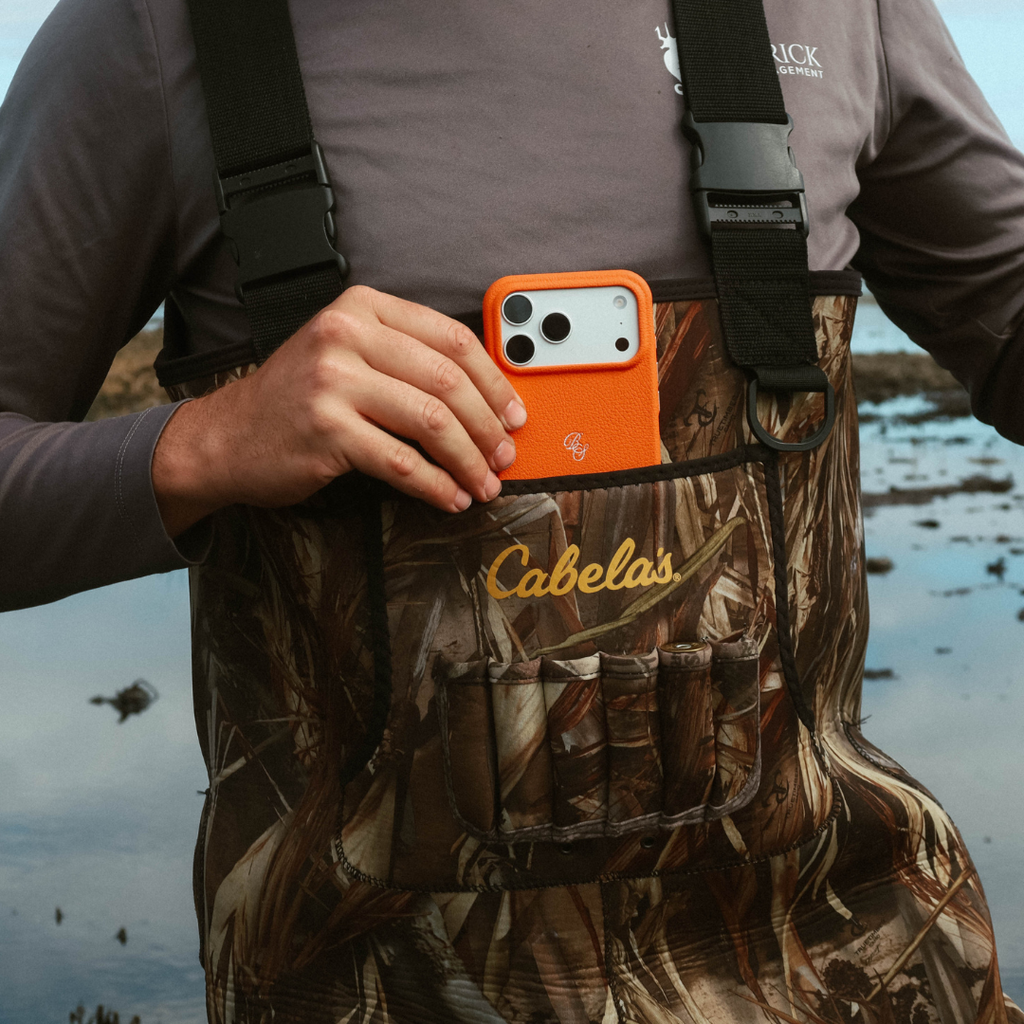 Person placing iPhone in hunter orange leather case into a camouflage wader chest pocket at a marshy outdoor setting