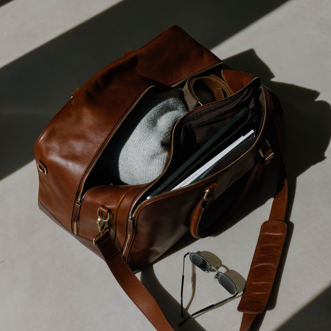 Brown leather duffel bag partially unzipped showing knit beanie, belt, papers and sunglasses beside it