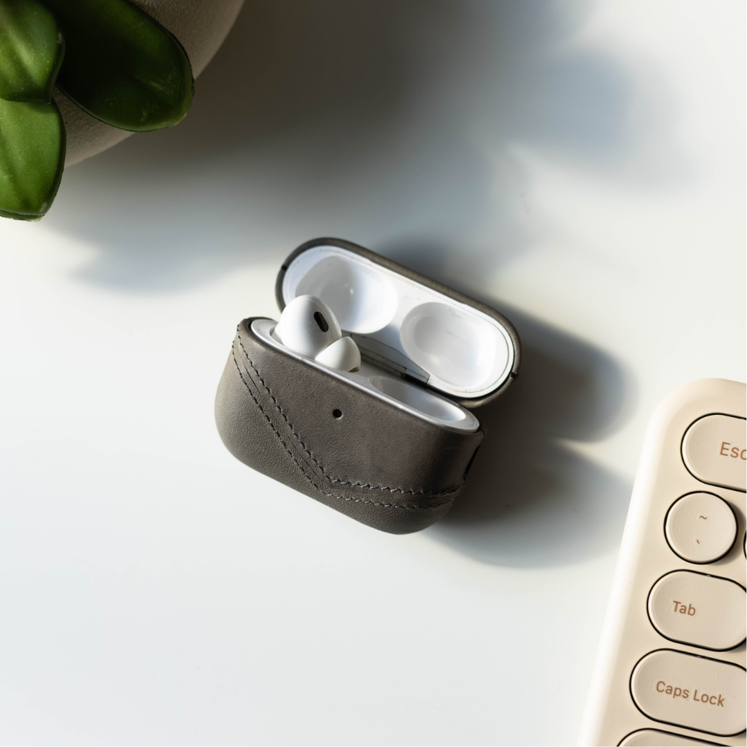 Slate leather AirPods case open with AirPods inside on white desk; keyboard keys: Esc, Tab, Caps Lock