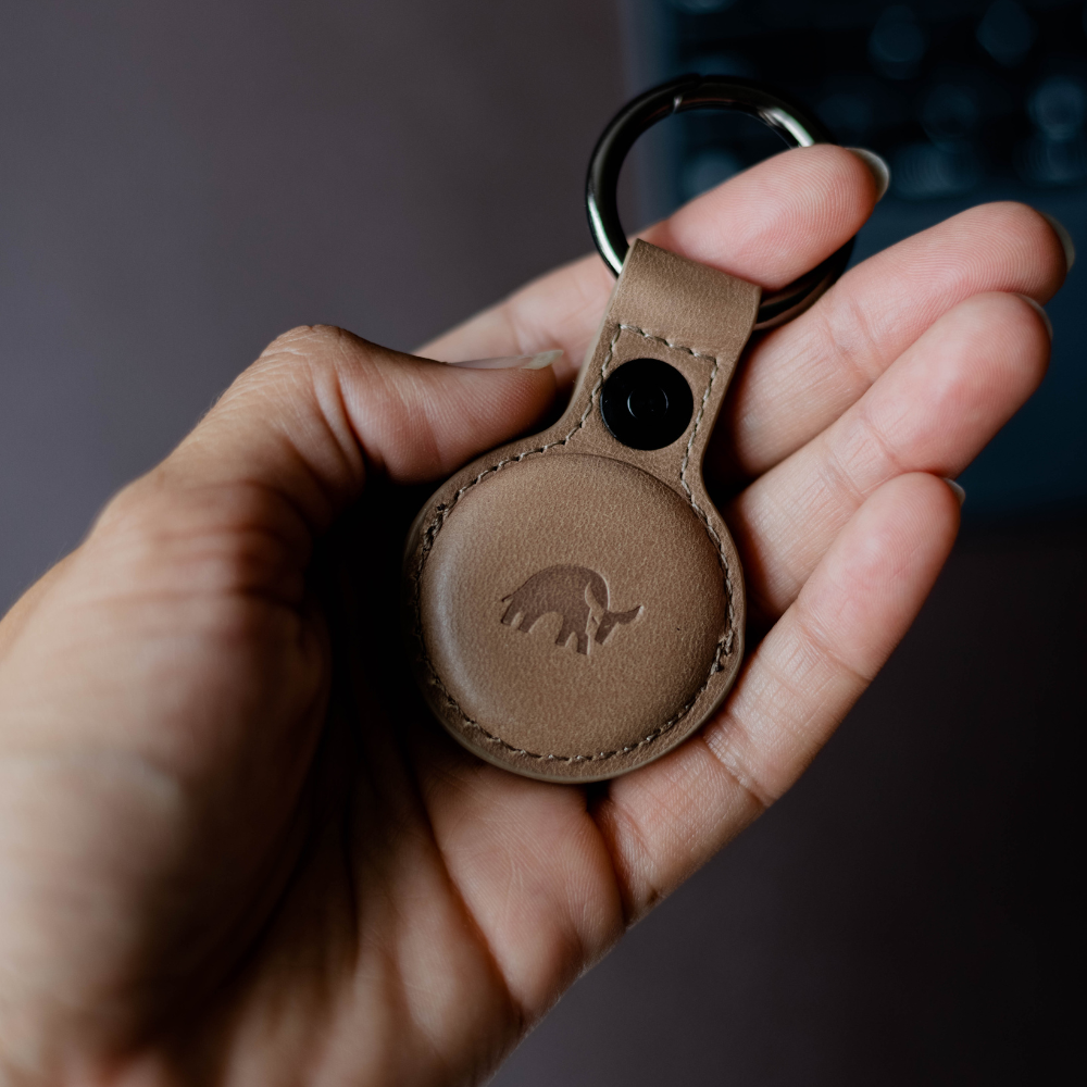A hand holding a BullTag for AirTag in Dune color, featuring a logo embossed on leather.