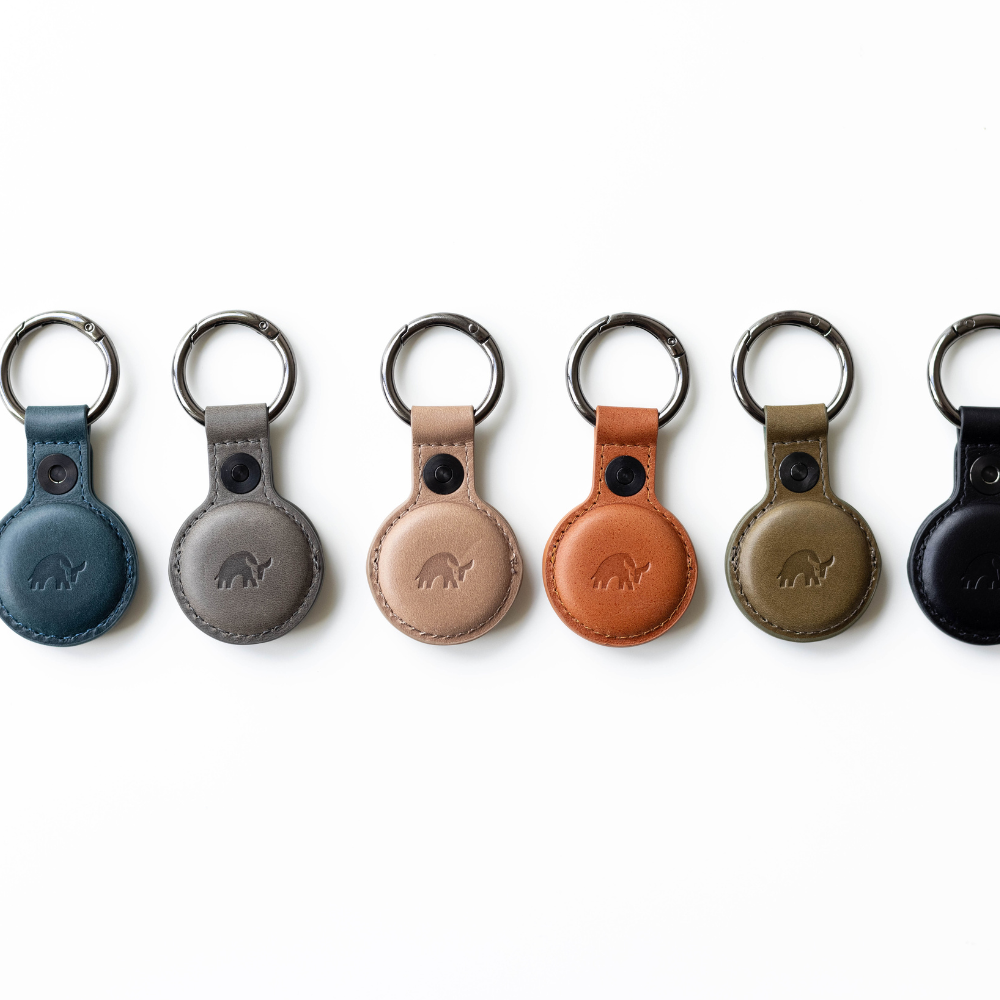 Six leather BullTag AirTag holders aligned horizontally in teal, gray, tan, caramel, olive, and black.