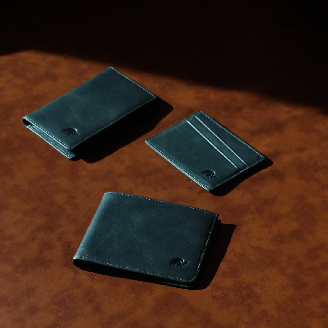 Three dark teal leather wallets and a slim card sleeve arranged on a brown leather surface, top-down view with soft shadows.