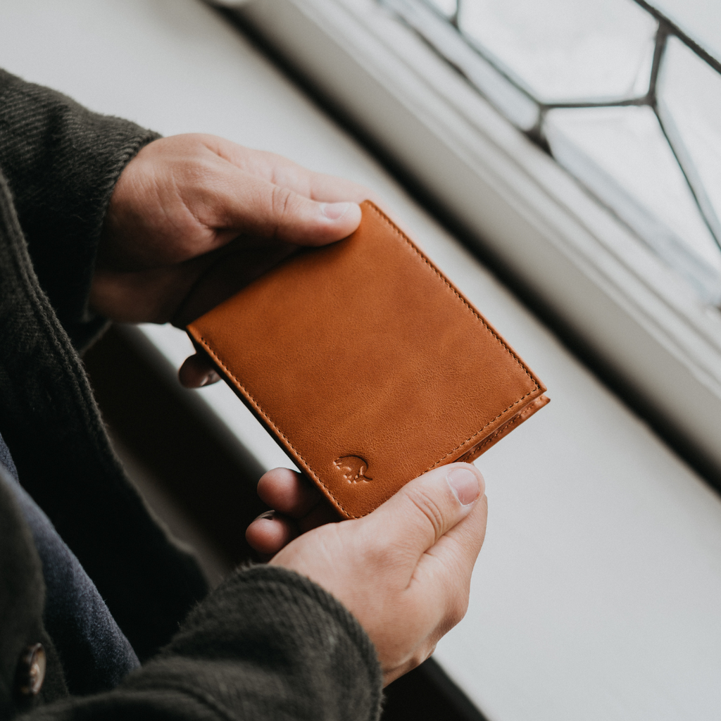 Brown sienna leather bifold wallet held in both hands near a window, small embossed logo on lower corner