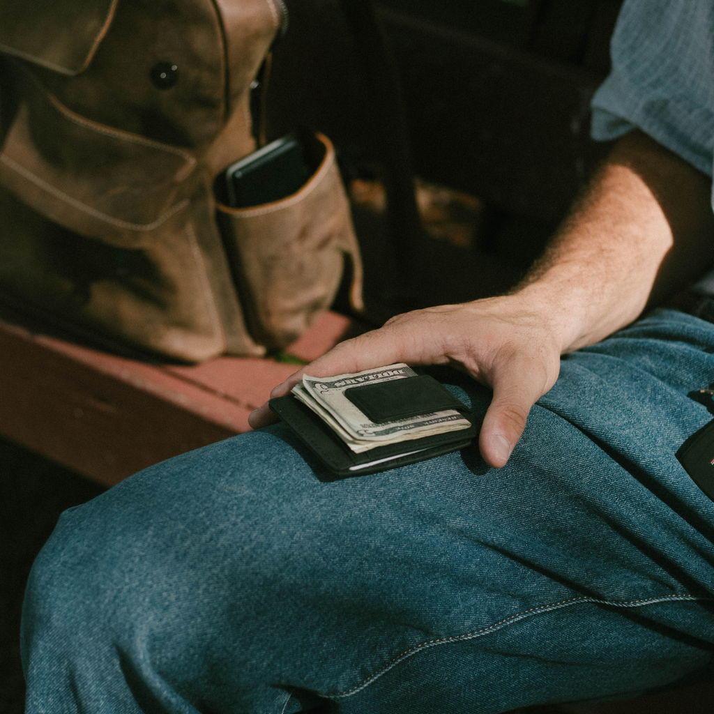 Hand resting on jean-clad thigh holding black money clip with folded U.S. bills