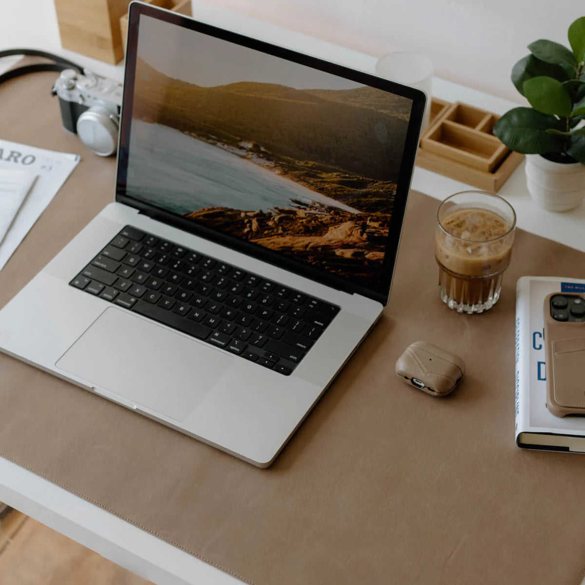 Tan leather desk mat with laptop, glass of iced coffee, camera, earbuds case, book and potted plant on white desk.